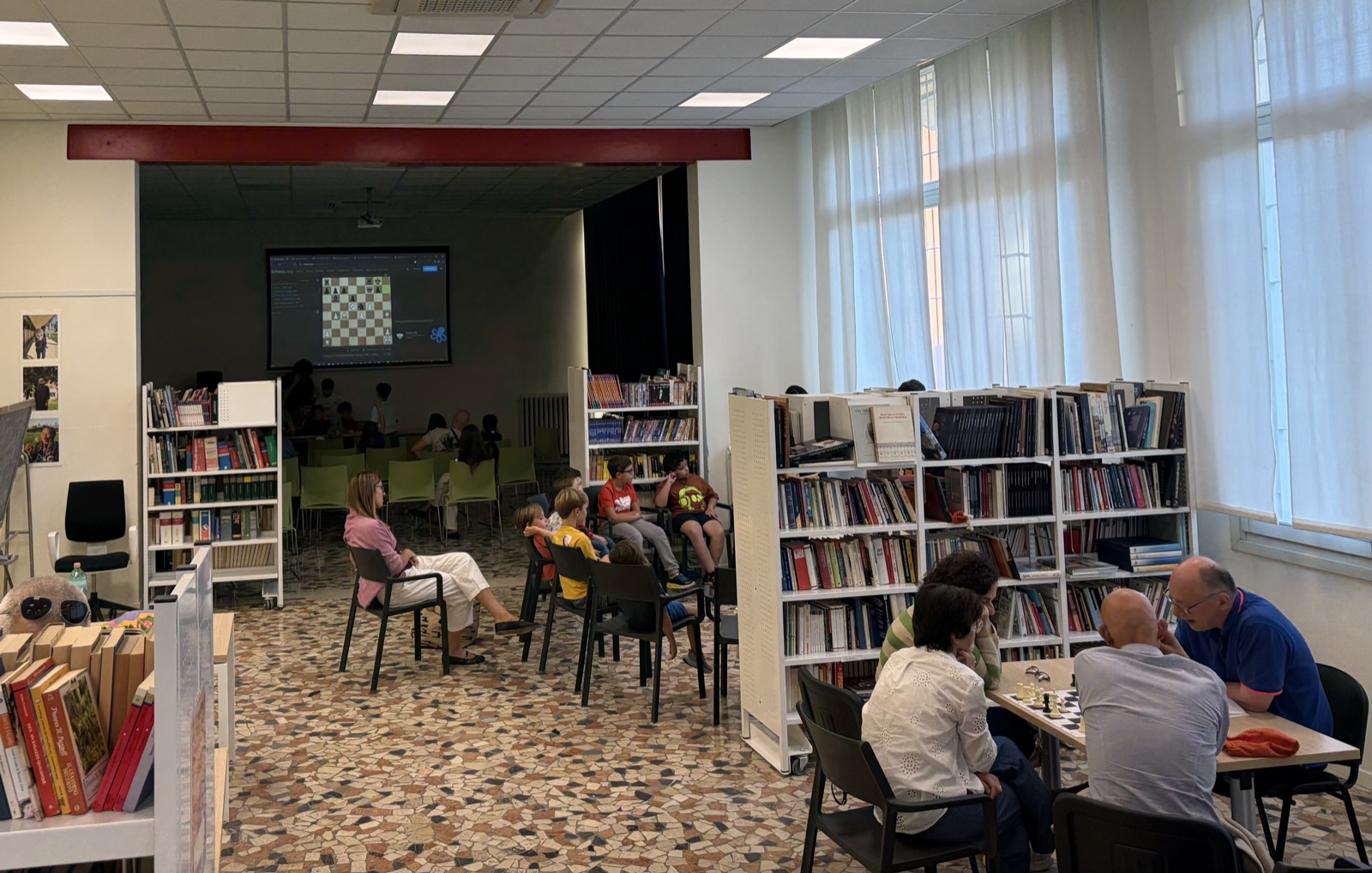 Lezione del corso con la scacchiera proiettata e soci che giocano tra le librerie della sede
